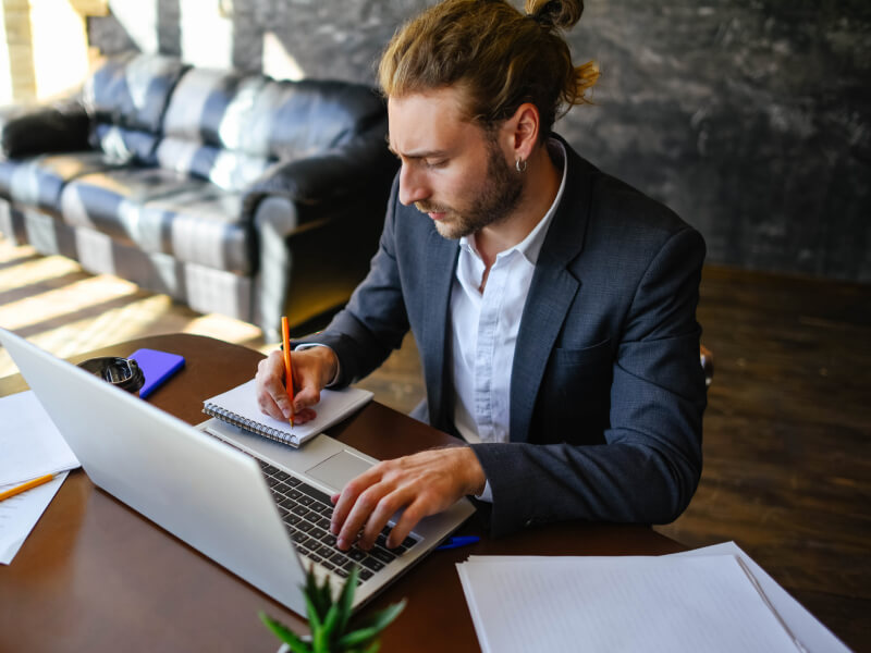 Man writing in his notebook while typing on the laptop